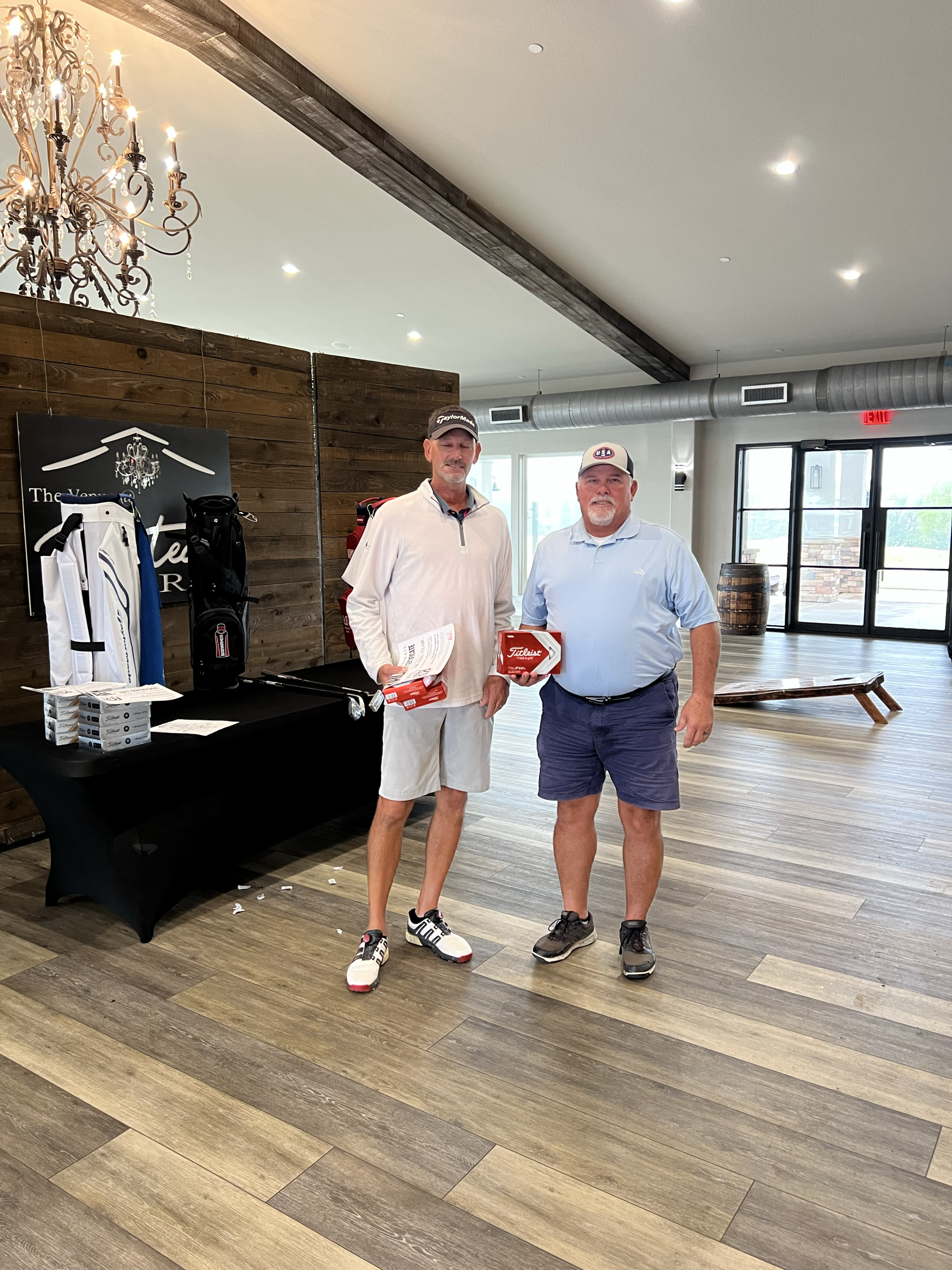 Two men holding trophies, indoors. One in white shirt, shorts, other in blue shirt, shorts.