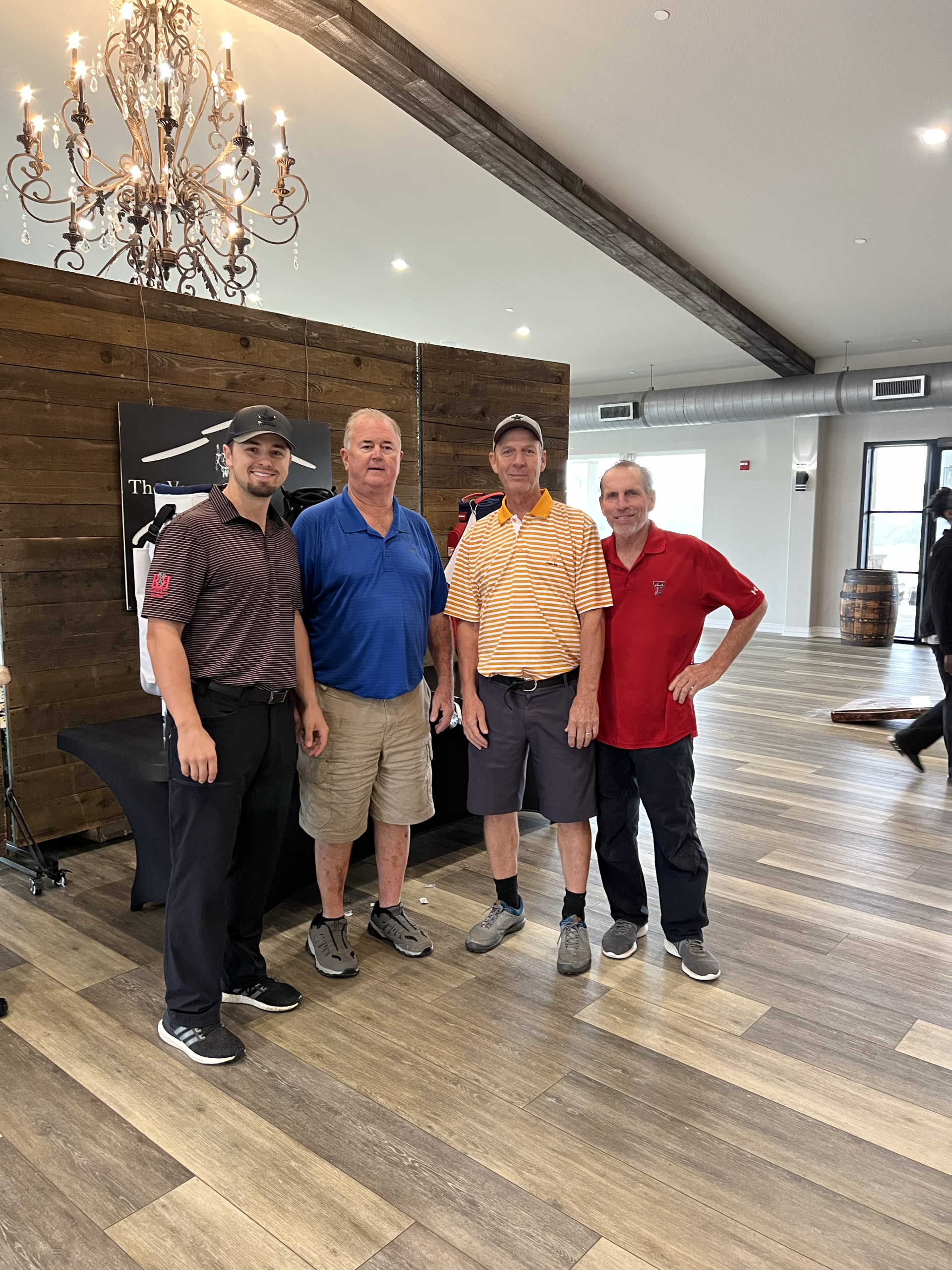 Four men pose inside, one with hand on hip. Wooden beams, chandelier and a barrell are visible.