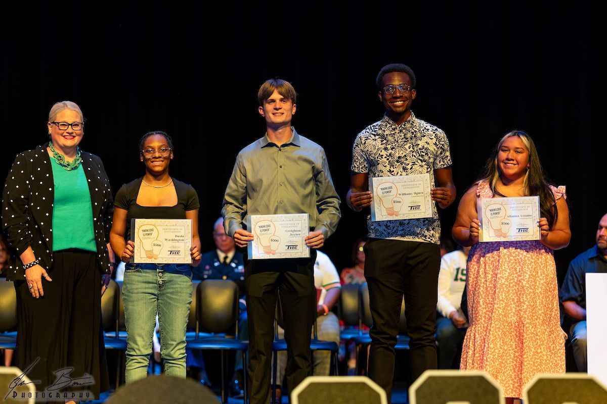 Five people on stage holding certificates, smiling. Woman in black blazer, three students, and a student in a floral shirt.