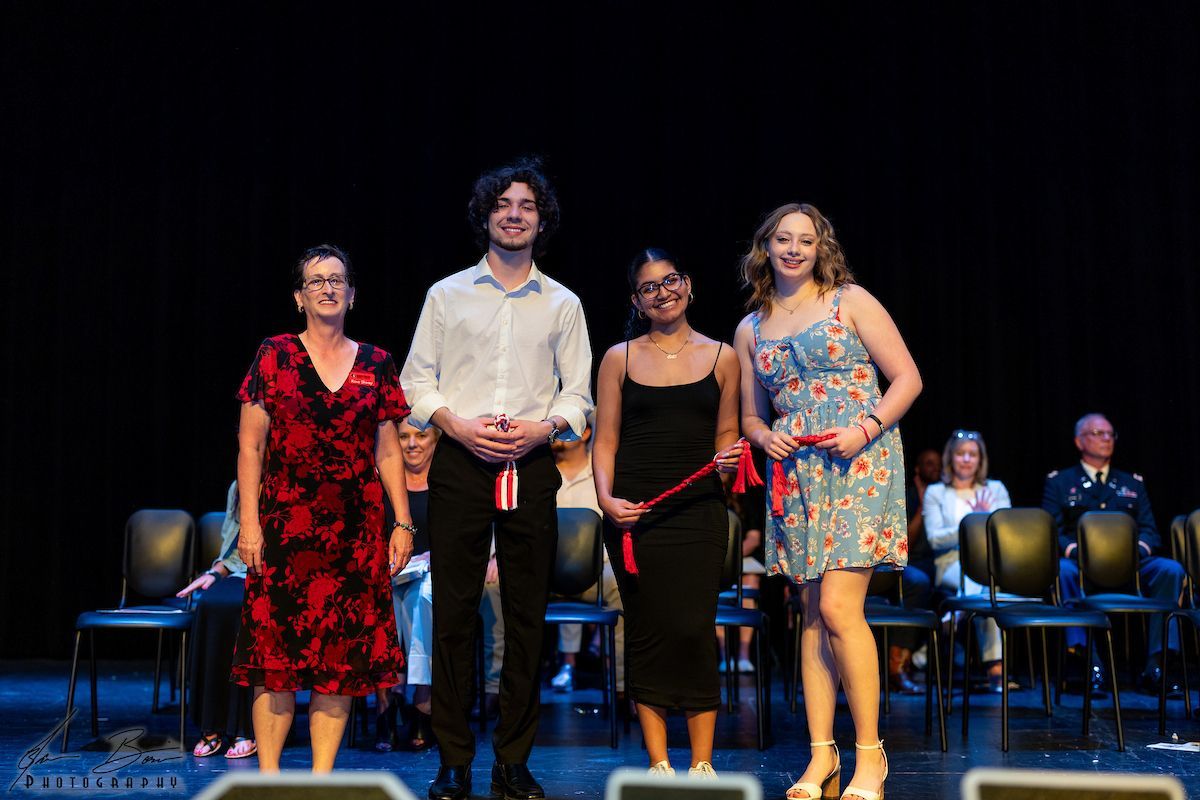 Four people on stage, holding red ribbons, smiling, with an audience seated behind them.