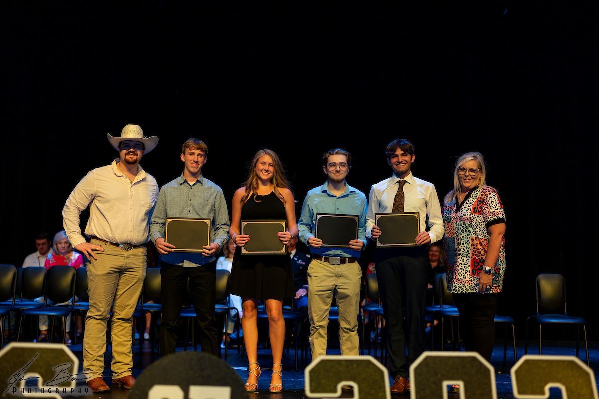 Group of six people on a stage, holding plaques. Two men wear cowboy hats. Background: black stage.