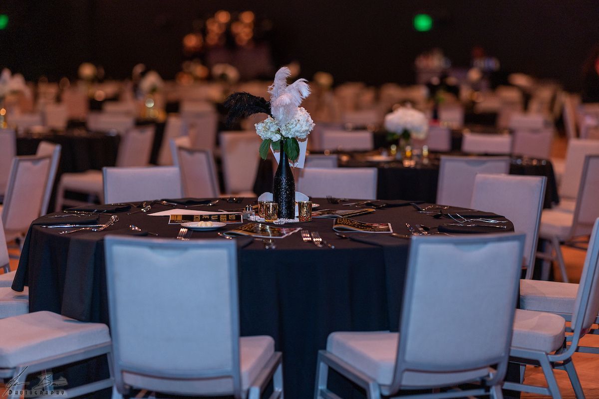 Round tables set for a formal event; black tablecloths, white chairs, floral centerpieces.