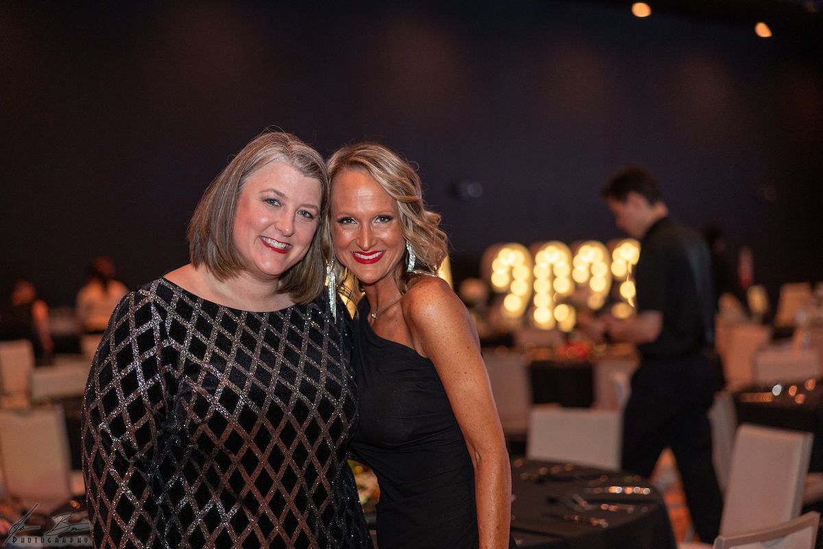 Two women smiling at a gala, one in a black sequined dress, one in a black one-shoulder dress. Large illuminated 2023 sign.