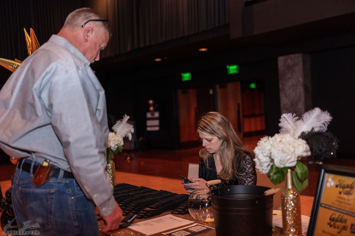 Man at registration table with woman, white flowers. Dark room, golden accents.