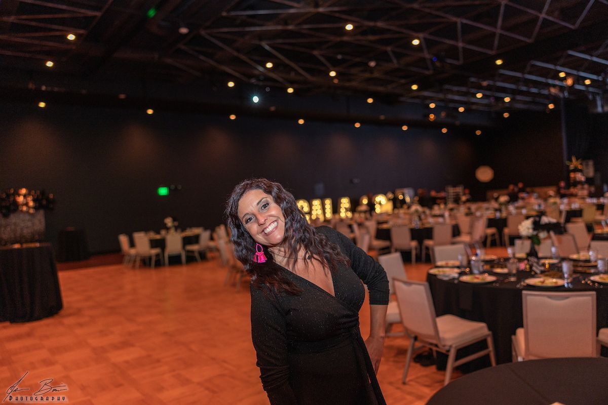 Woman smiles in a ballroom with tables set for a gala; black attire, pink earrings.