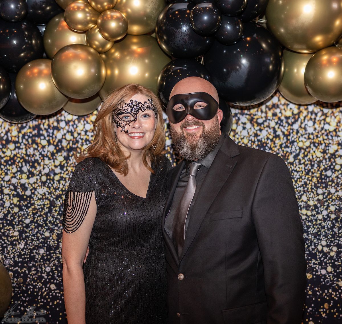 Couple in masquerade masks pose in front of balloons and glitter backdrop. Woman wears black dress. Man in suit.
