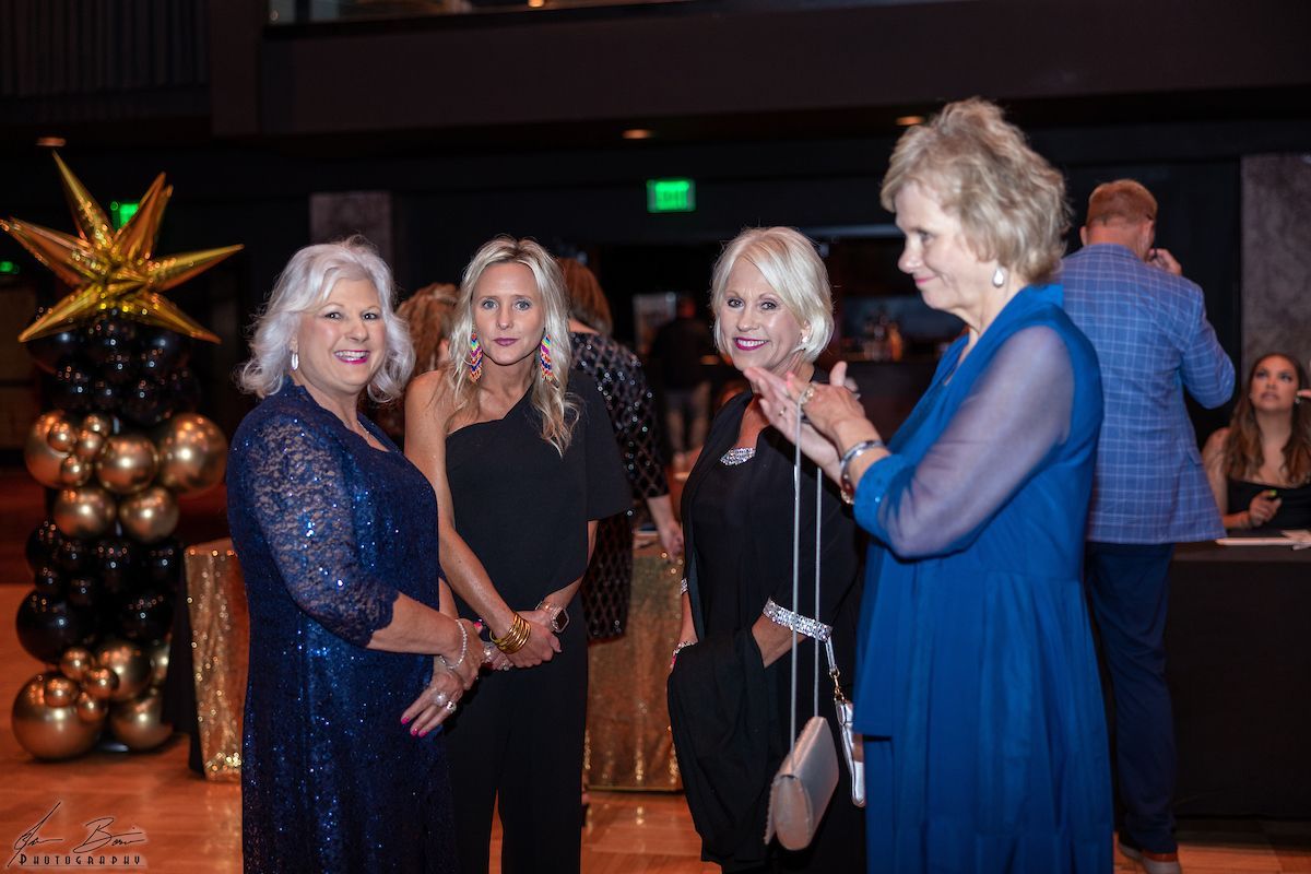 Four women at an event, one in blue gestures, others smiling. Gold and black balloons in background.