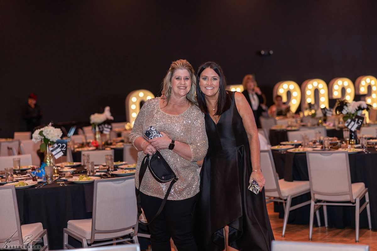 Two women smiling at a gala. One in a gold sequin top, the other in a black dress, posing near tables and decor.