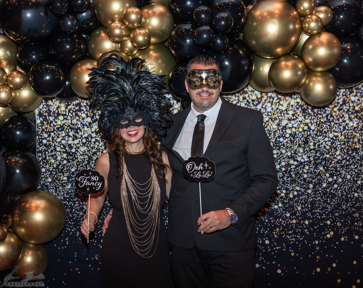 Couple in masks at a party, posing with props in front of gold/black balloon arch backdrop.