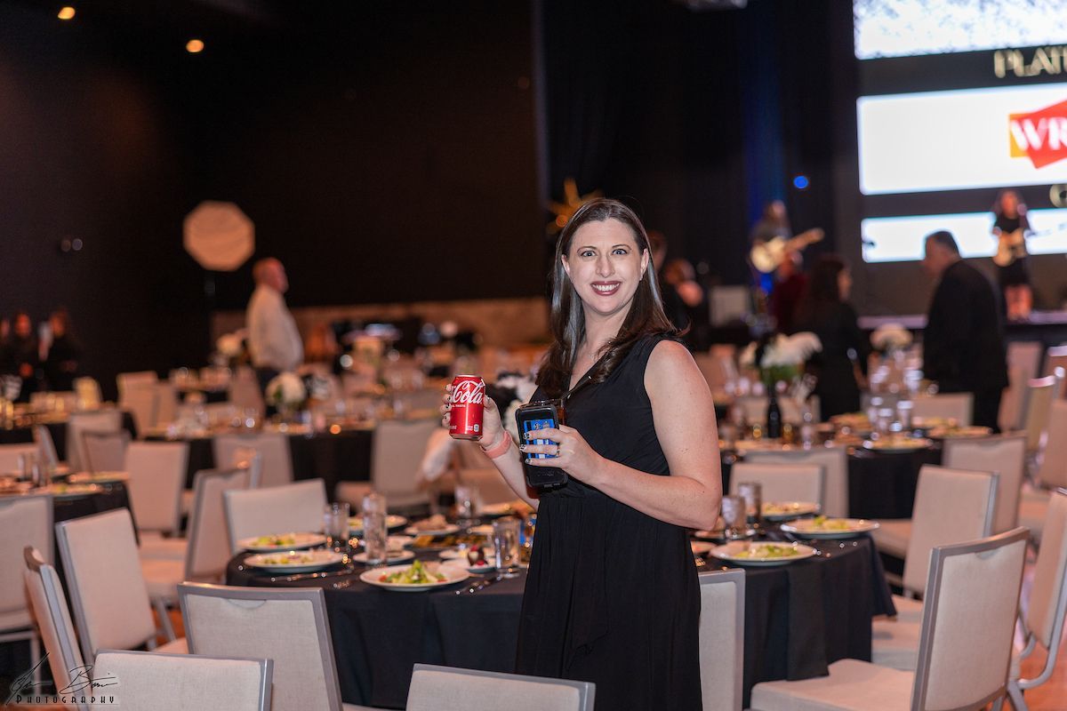 Woman in black dress at an event, holding drinks, smiling, tables set, band in background.