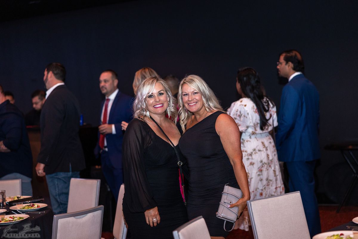 Two women smile in black dresses at an event with other formally dressed people.