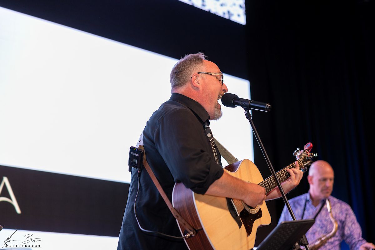 Man singing and playing acoustic guitar on stage, another man with a saxophone in background.