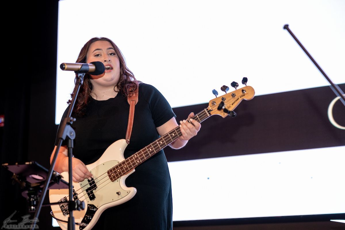 Woman singing and playing a white bass guitar onstage; white screen in the background.