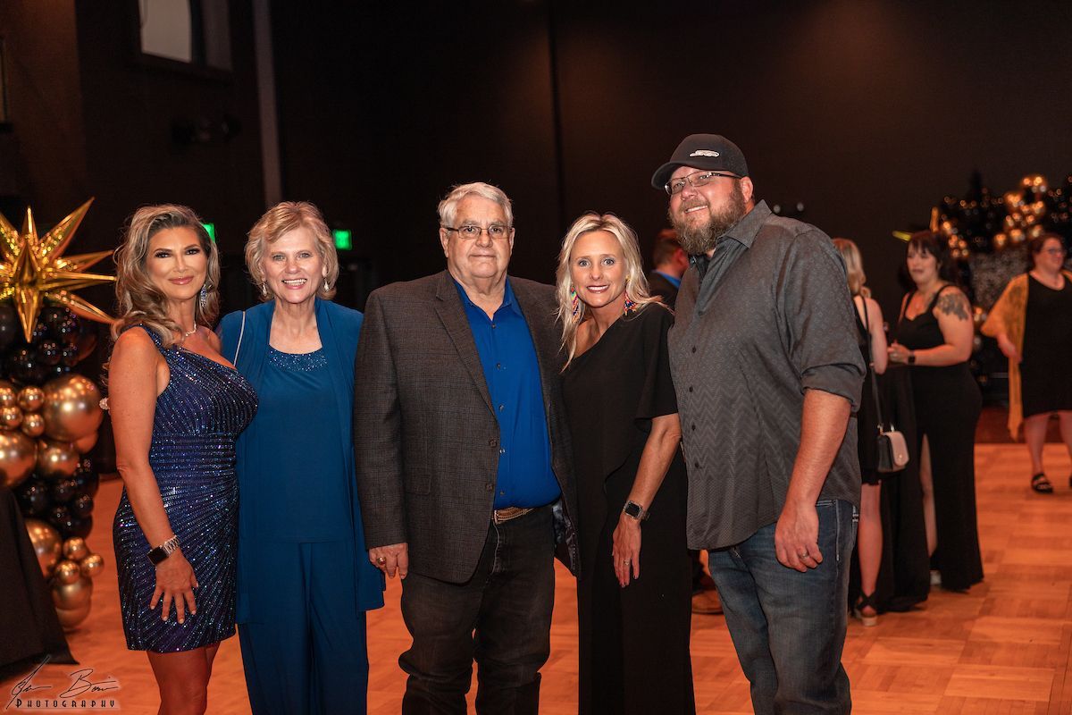 Five people posing at an event with gold and black balloons. Men in a suit and hat. Women in dresses.