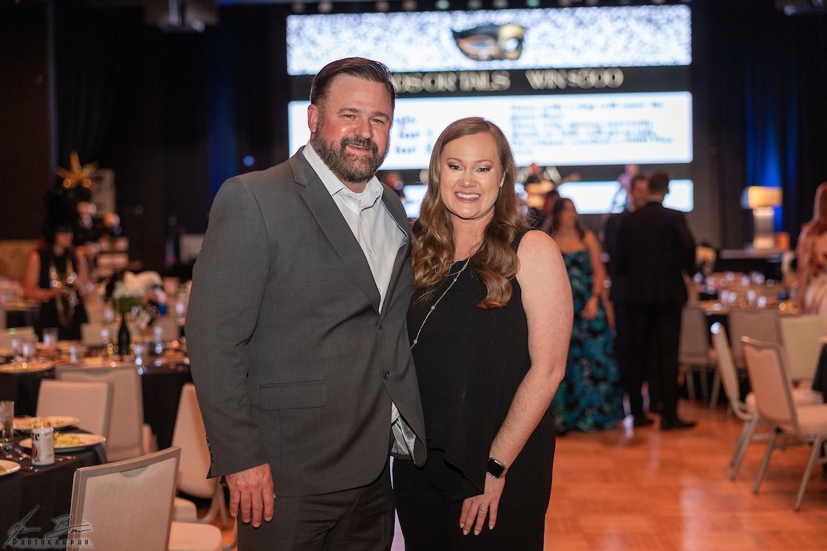 Man in grey suit and woman in black dress pose at a gala, smiling. Tables and decor are in the background.