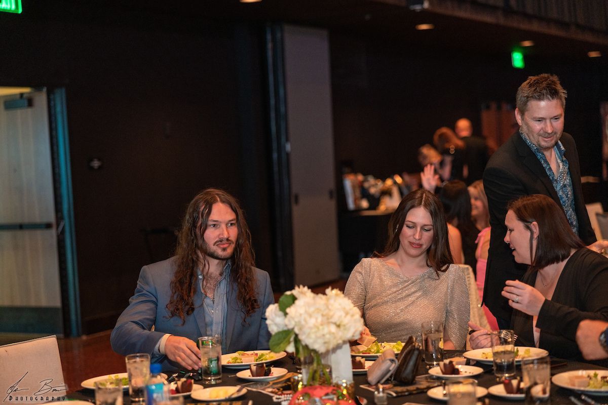 People seated at a dinner table; man with long hair looks at the camera. Others converse nearby. Dark room.