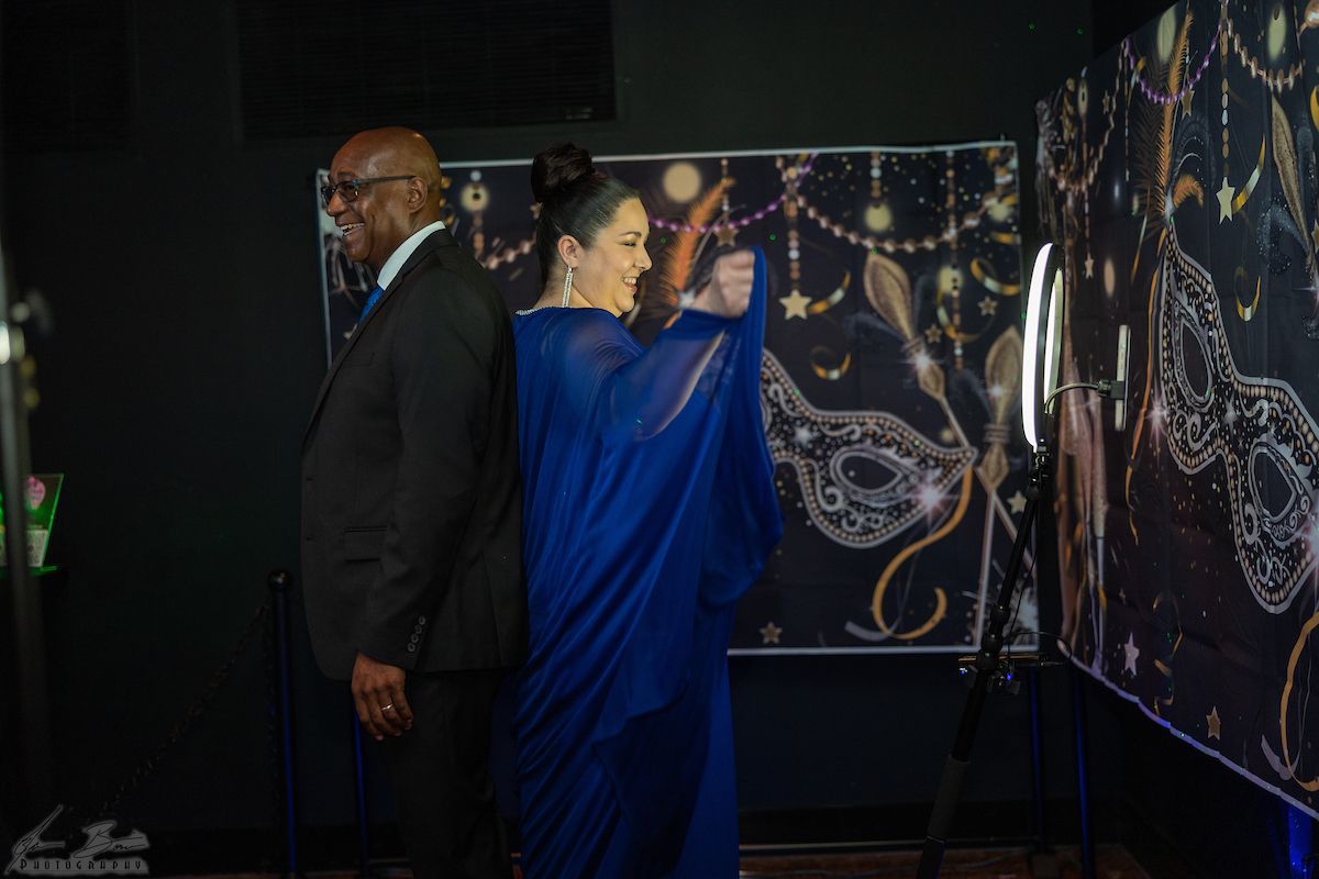 Man and woman at photo booth, woman poses in blue dress, man in suit. Black backdrop, ornate design.
