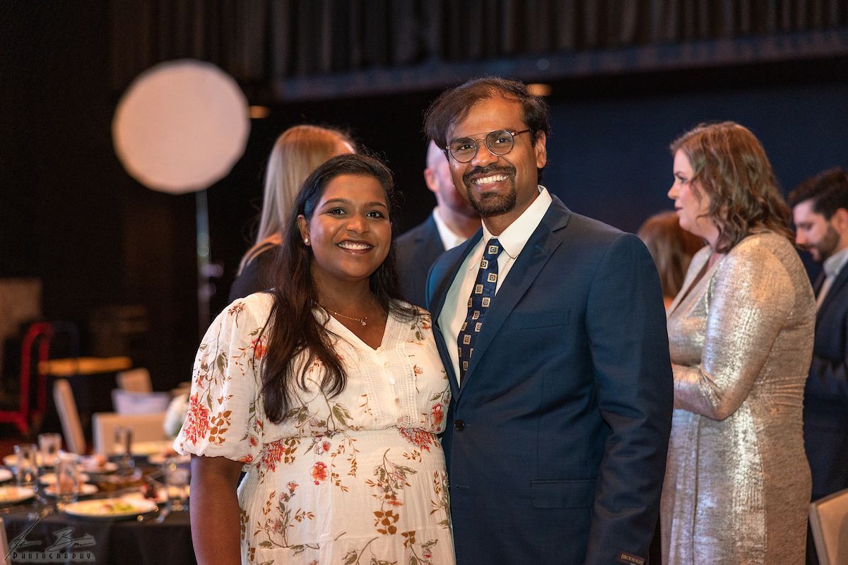 Smiling couple at an event, woman in floral dress, man in suit, others in background.