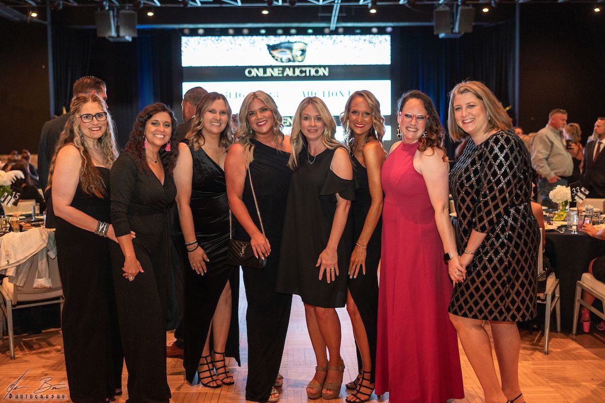 Group of women at an event. They are posing in various black dresses, smiles, on a dance floor.