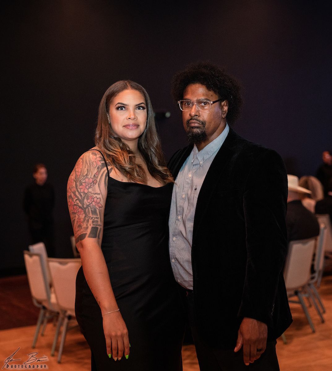 Woman in black dress and man in blazer pose together. Indoor setting with audience.