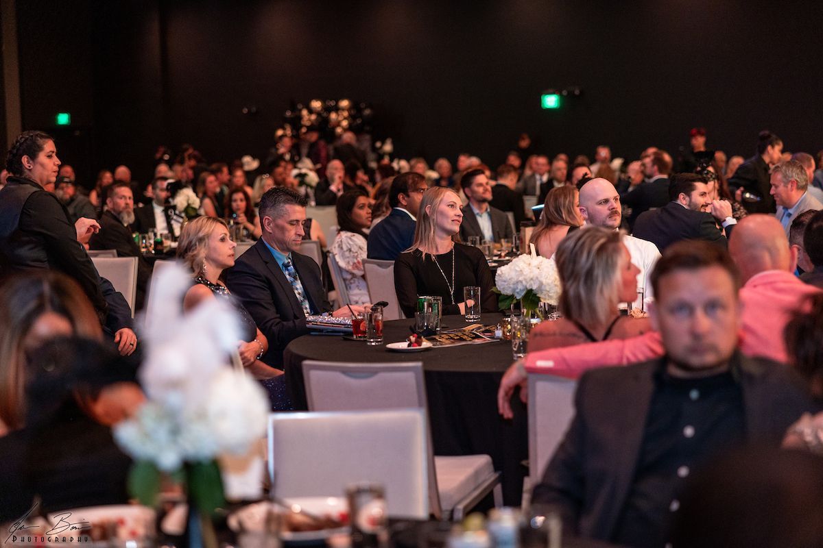 People seated at tables in a large, dimly lit ballroom. Many are looking forward, likely at a stage or presentation.
