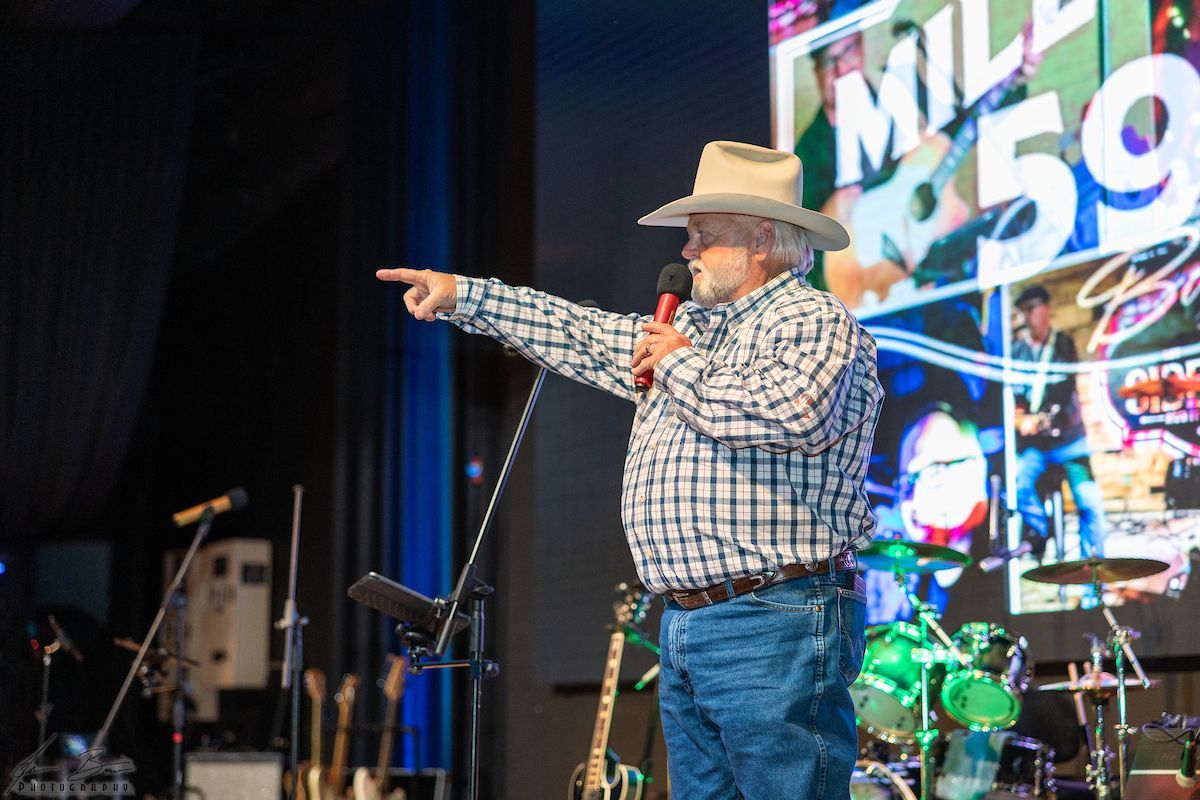 Man in cowboy hat points to the side while speaking on stage; band equipment and screen in background.