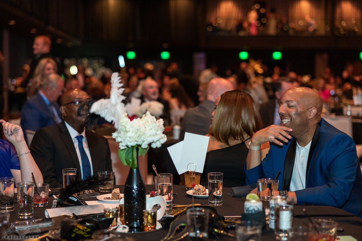 Guests at a formal event seated at tables, laughing, socializing. Dark setting, table decorations, and people in formal attire.