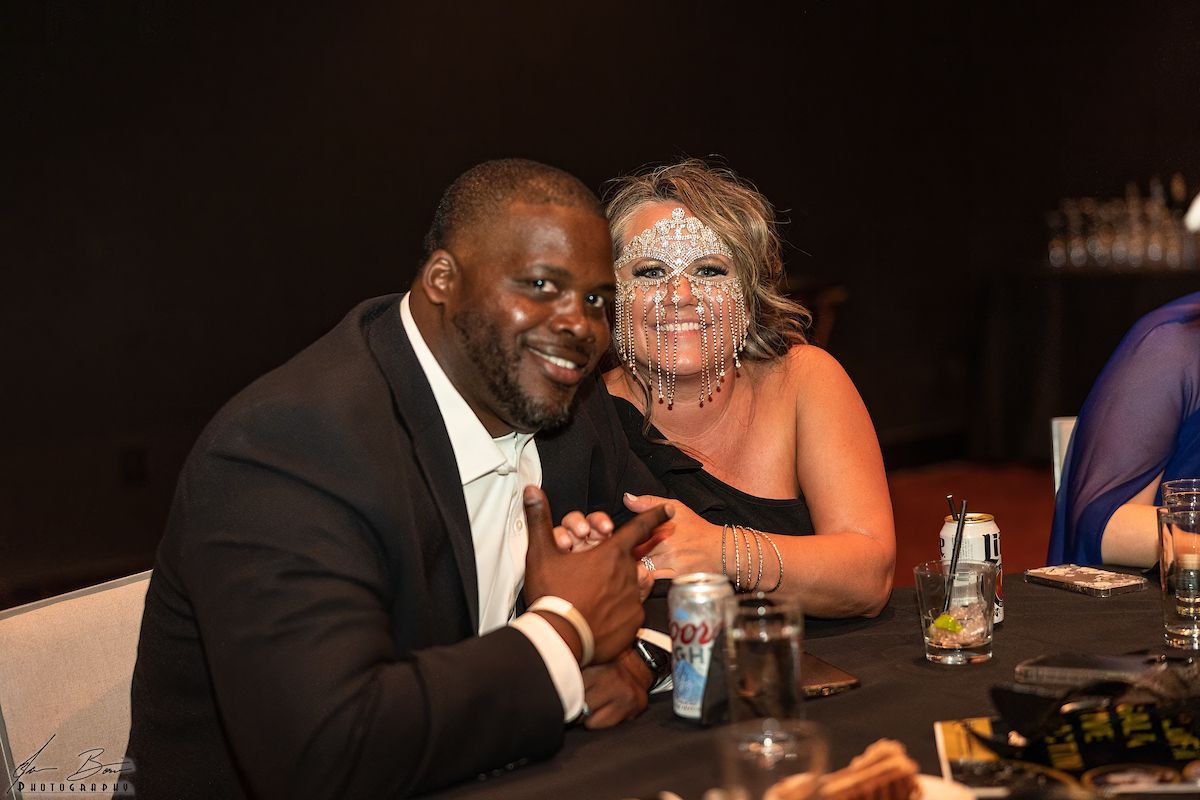 Man and woman at a table, smiling. Woman has icing on her face. Man is wearing a suit.