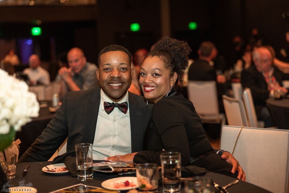 Couple smiling at a formal event, man in suit and bowtie, woman in black. Indoor setting with tables and guests.