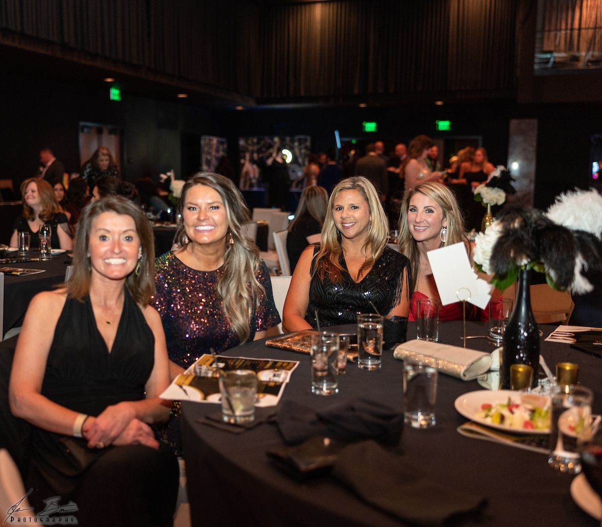 Four women smiling at a formal event, seated at a table. Black tablecloth, drinks, and decorations.