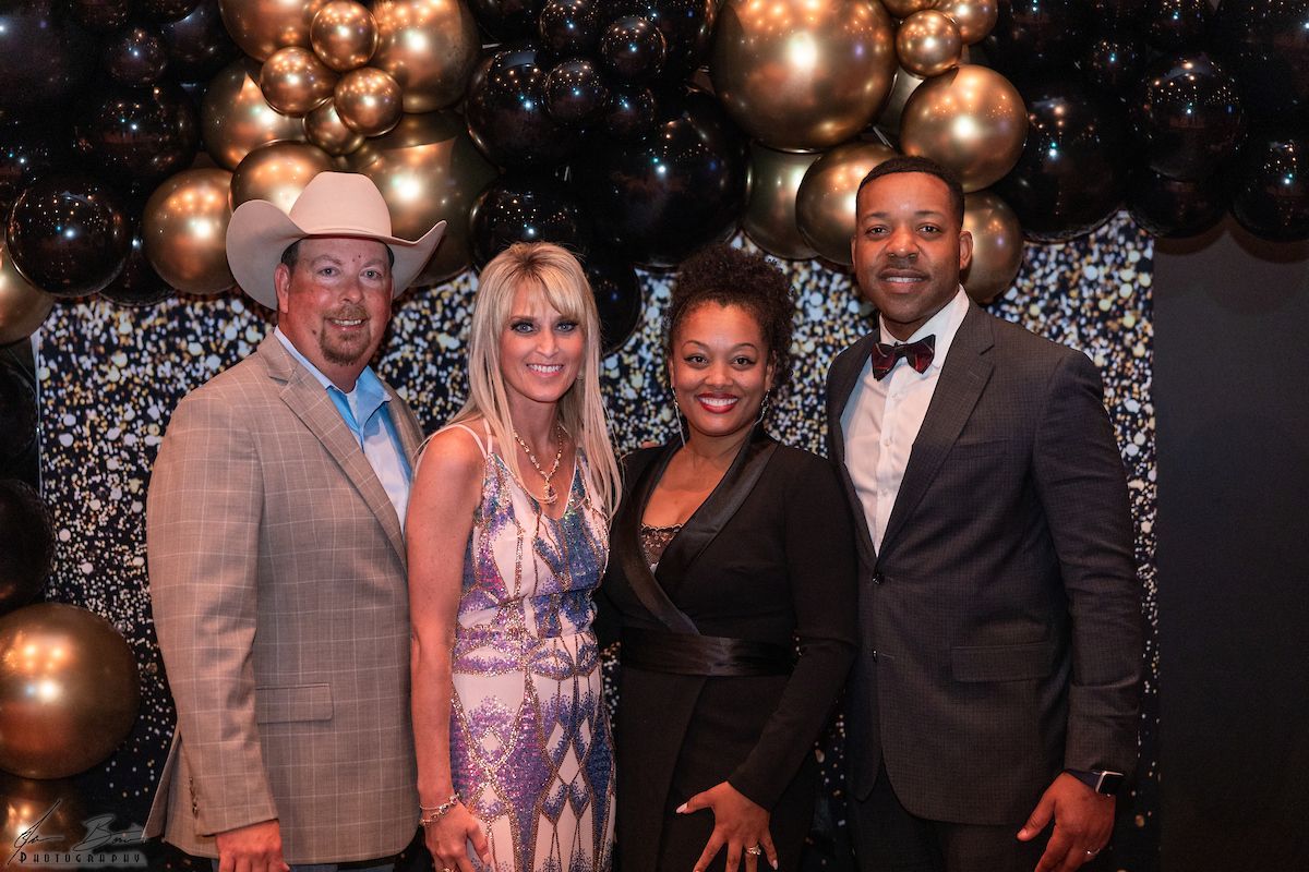 Four people smiling at a formal event with a gold and black balloon backdrop.