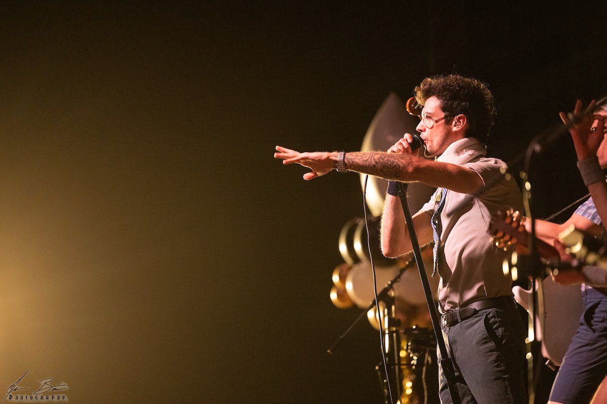 Man singing, arm raised, with brass instruments and a performer on stage.