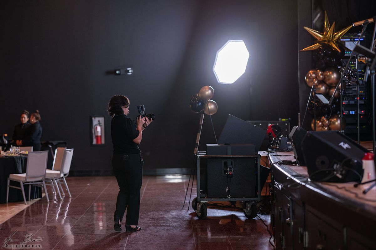 A person filming an event with camera, bright overhead light, black wall, and gold balloons.