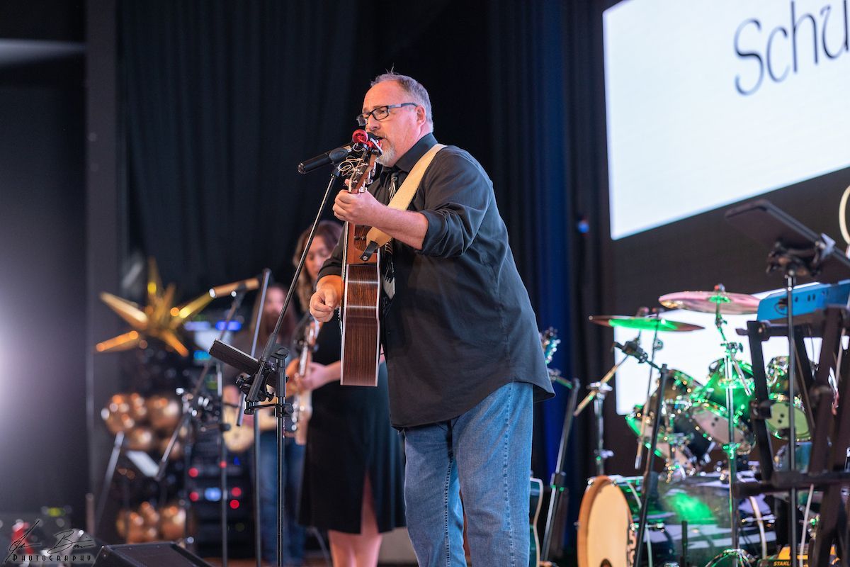 Man singing and playing guitar on stage with band and audience.