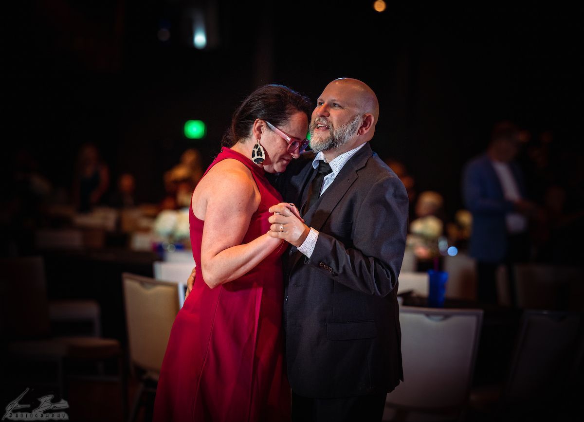 Couple dancing at an event; woman in red dress, man in suit, smiling, holding hands in front of a blurred background.