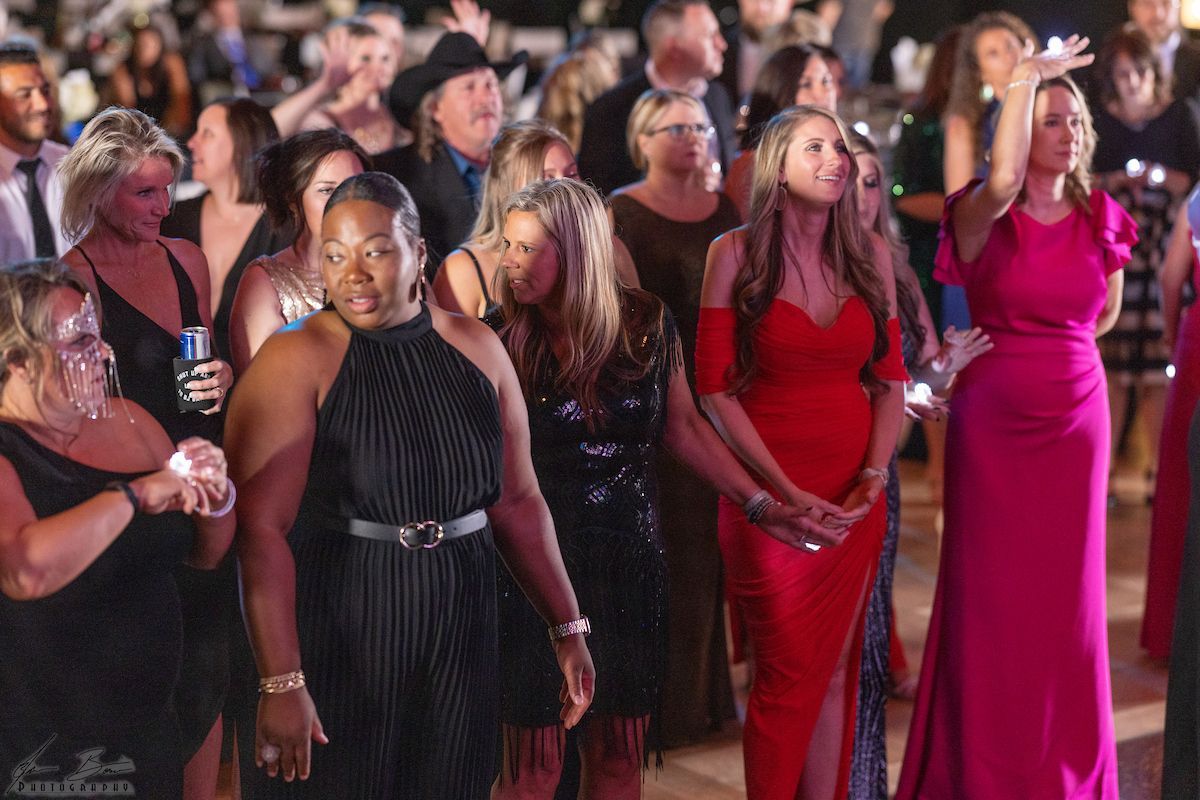 A crowd of formally dressed people at an event; women in gowns, some with raised hands, dancing.