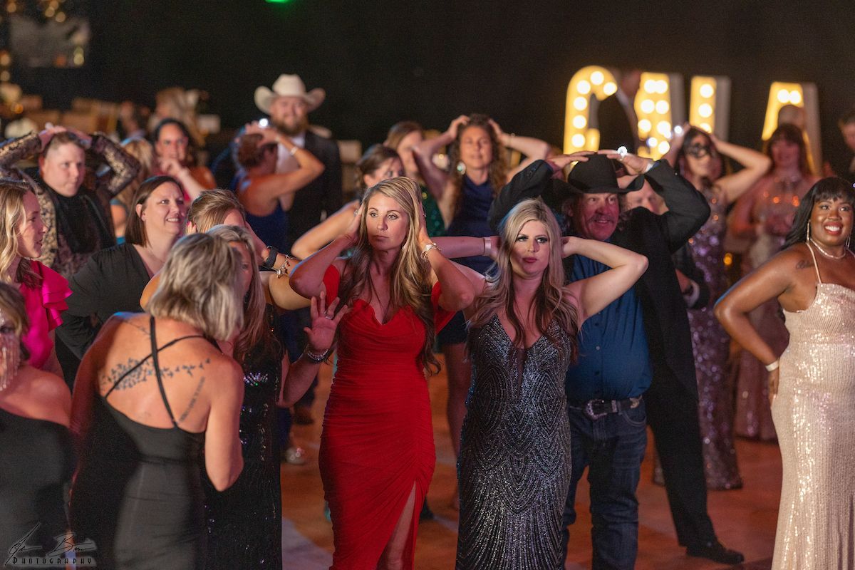 Group of people in formal wear dancing, raising hands. Lights spell 