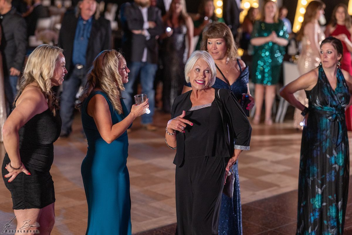 Group of women at a party, chatting and gesturing on a dance floor. Some wear evening gowns.