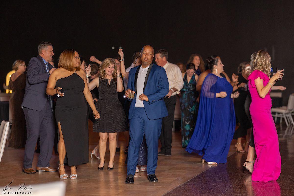 People dancing at an event in a large room, with diverse outfits, lively expressions, and a dark backdrop.
