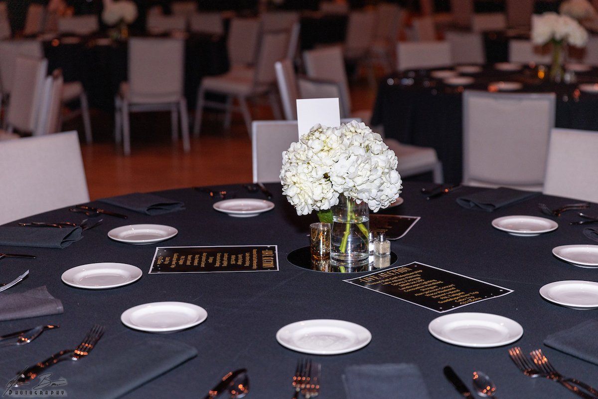Round tables set for a formal event, with white flowers, black tablecloths, and place settings.