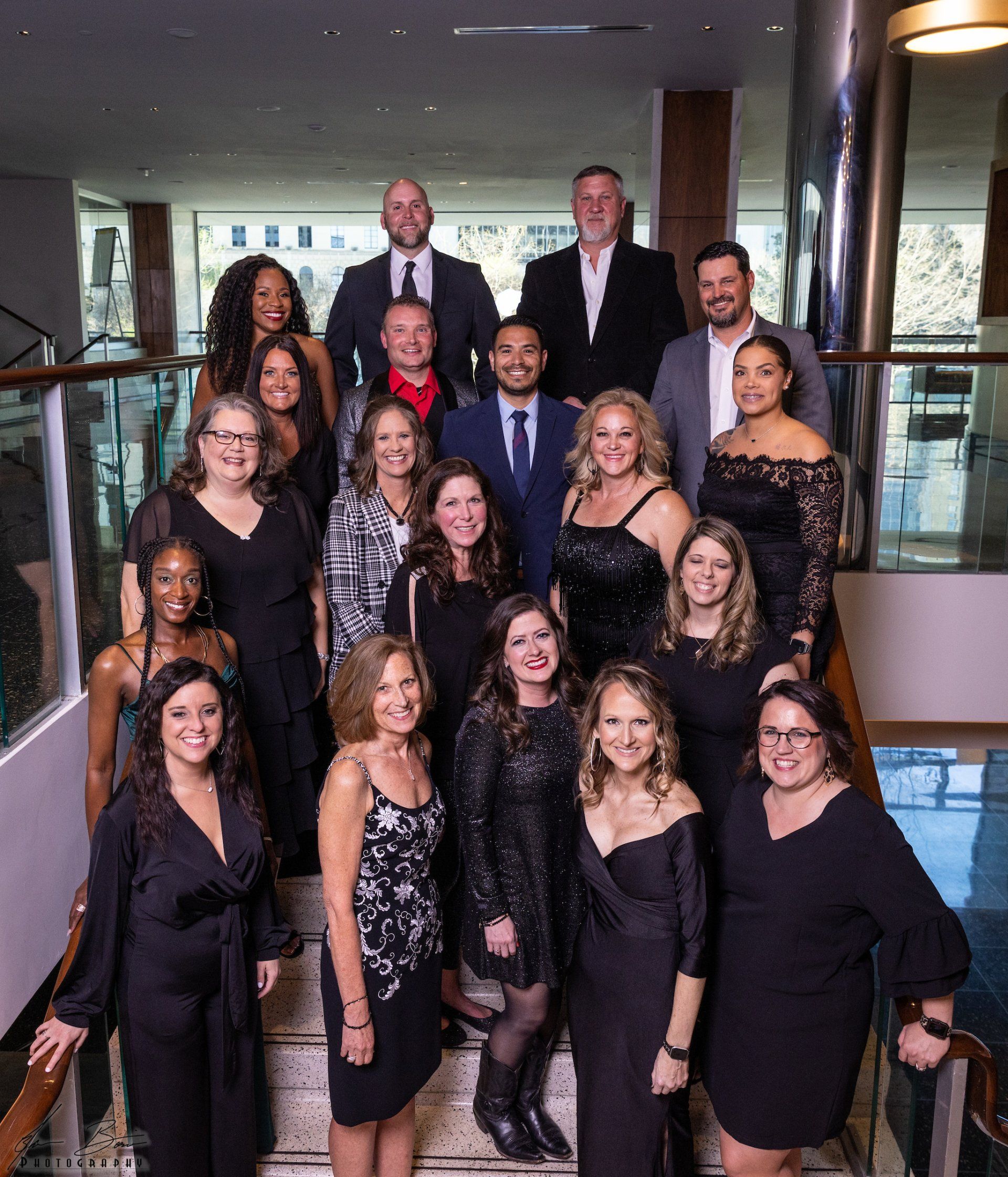 Group of people, mostly women, in formal wear, posing on steps indoors.