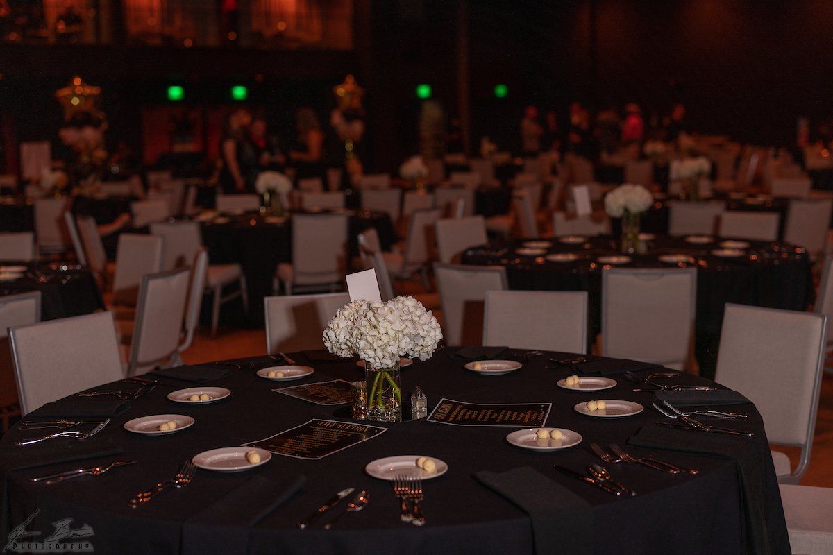 Formal event: round tables with black tablecloths, white chairs, centerpieces, guests in background.