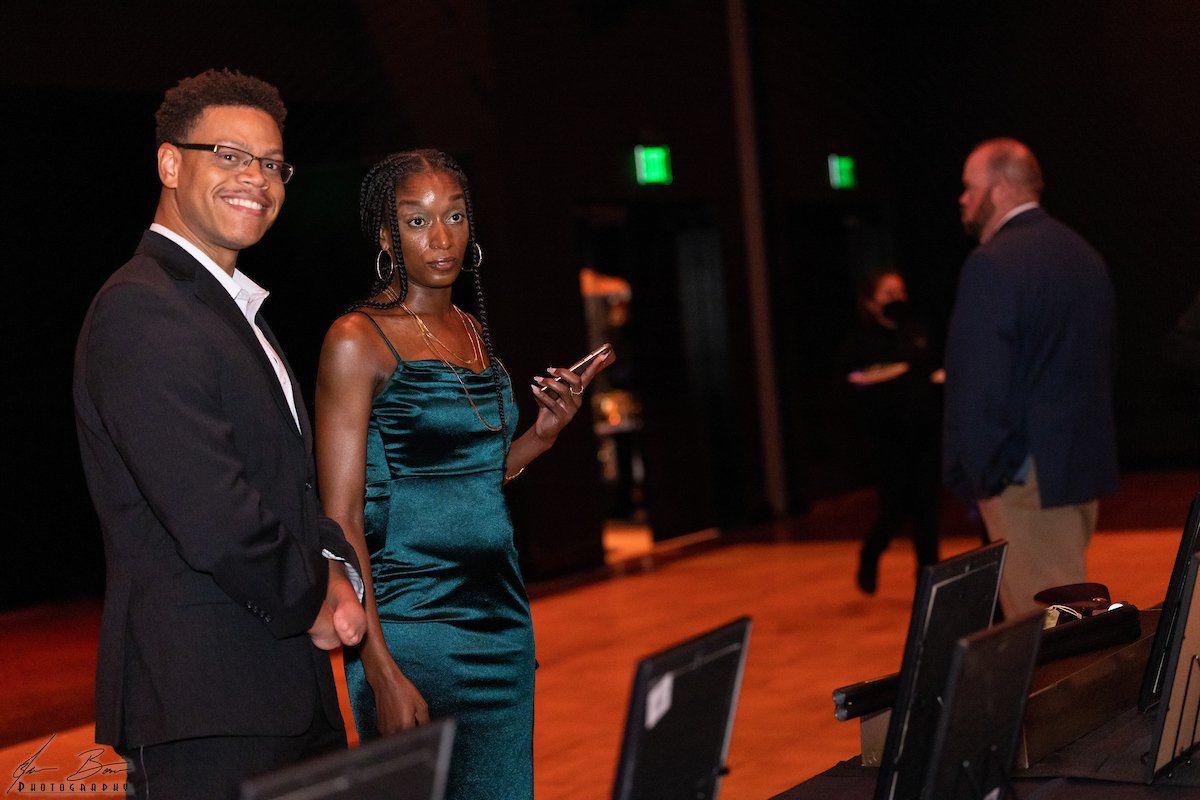 Two people in formal attire at an event. Woman in green dress gestures. Man in suit smiles. Dark stage setting.