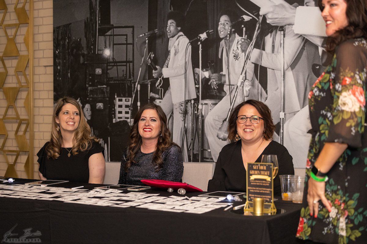 Women at a registration table smiling, with a band mural backdrop.
