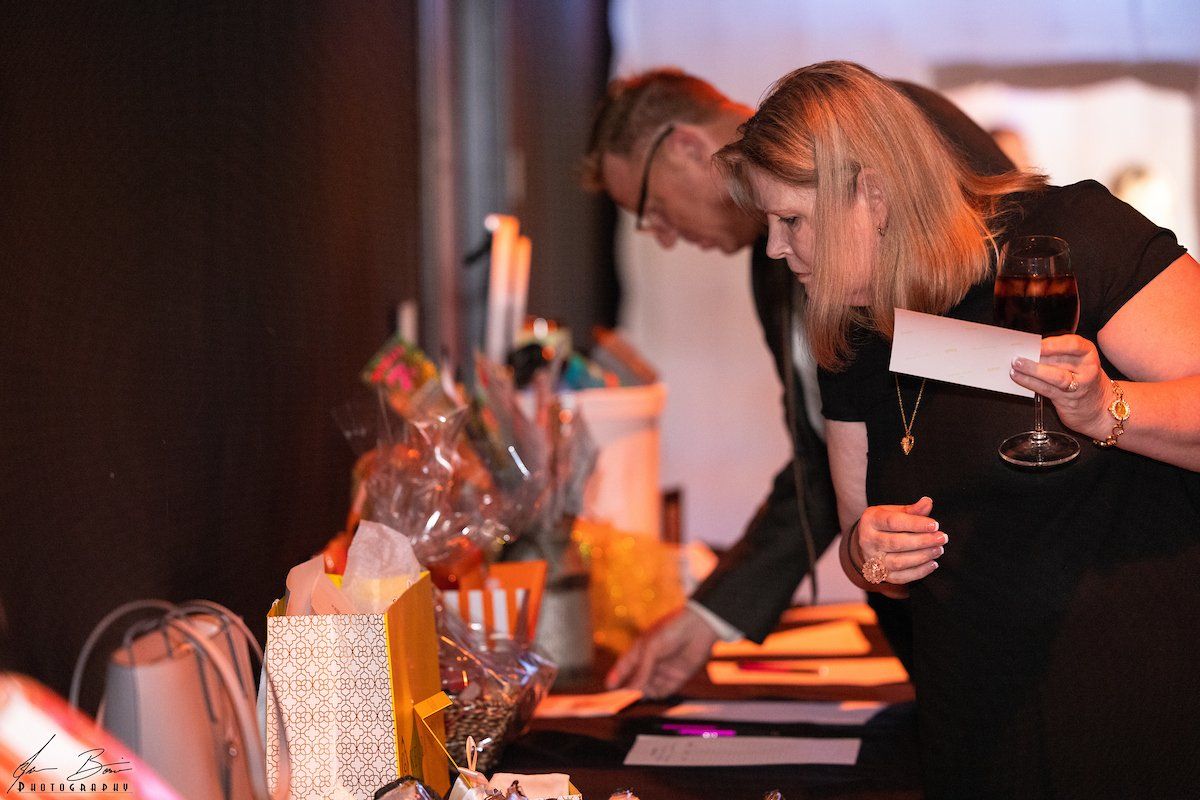 A man and woman looking at items on a table, likely at an auction or event. The woman holds a wine glass.