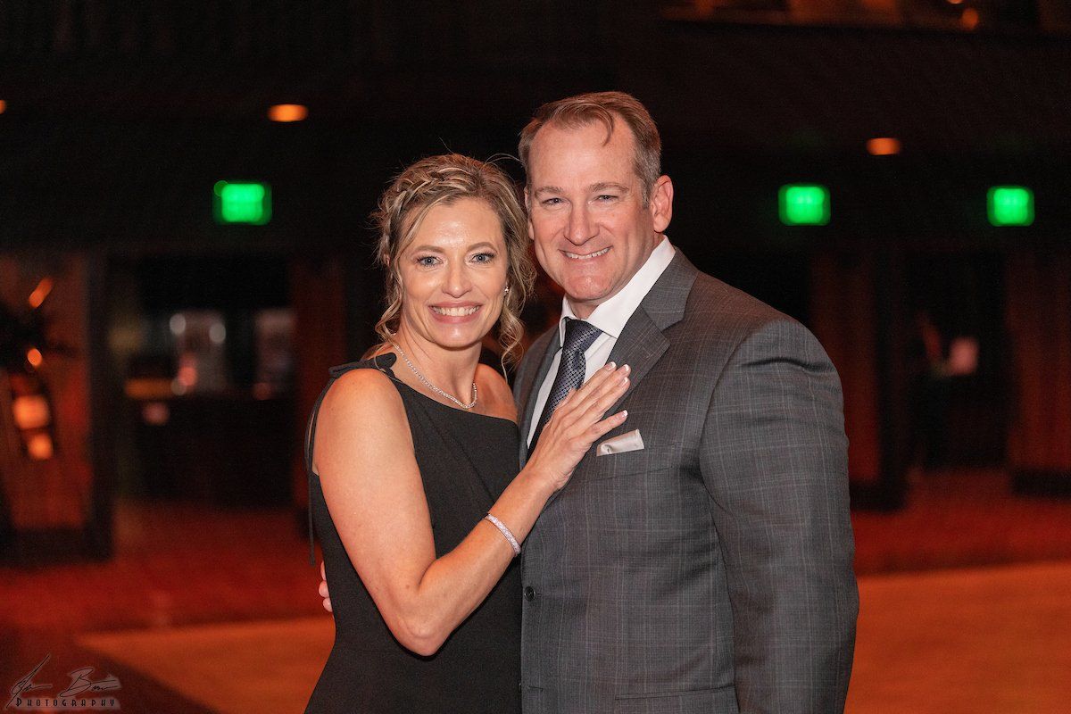 Couple in formal attire, woman in black dress, man in suit, smiling, standing in a dim, elegant setting.