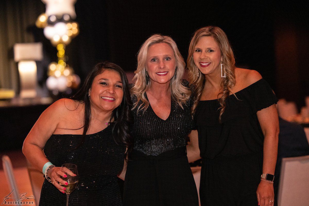 Three women in black dresses smile and pose together indoors; festive decorations in background.
