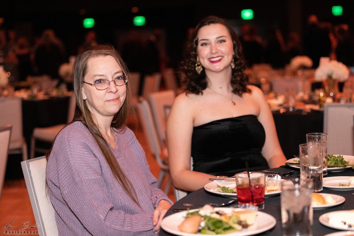 Two women at a formal event. One in glasses and a lilac sweater, the other in a black strapless dress.
