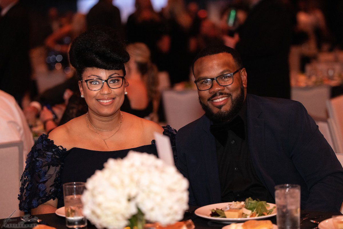 A smiling couple at a formal event, woman in a navy dress, man in a suit, seated at a table.