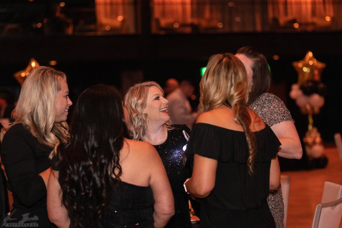 Five women in evening attire laughing at an event, with star-shaped balloons in the background.